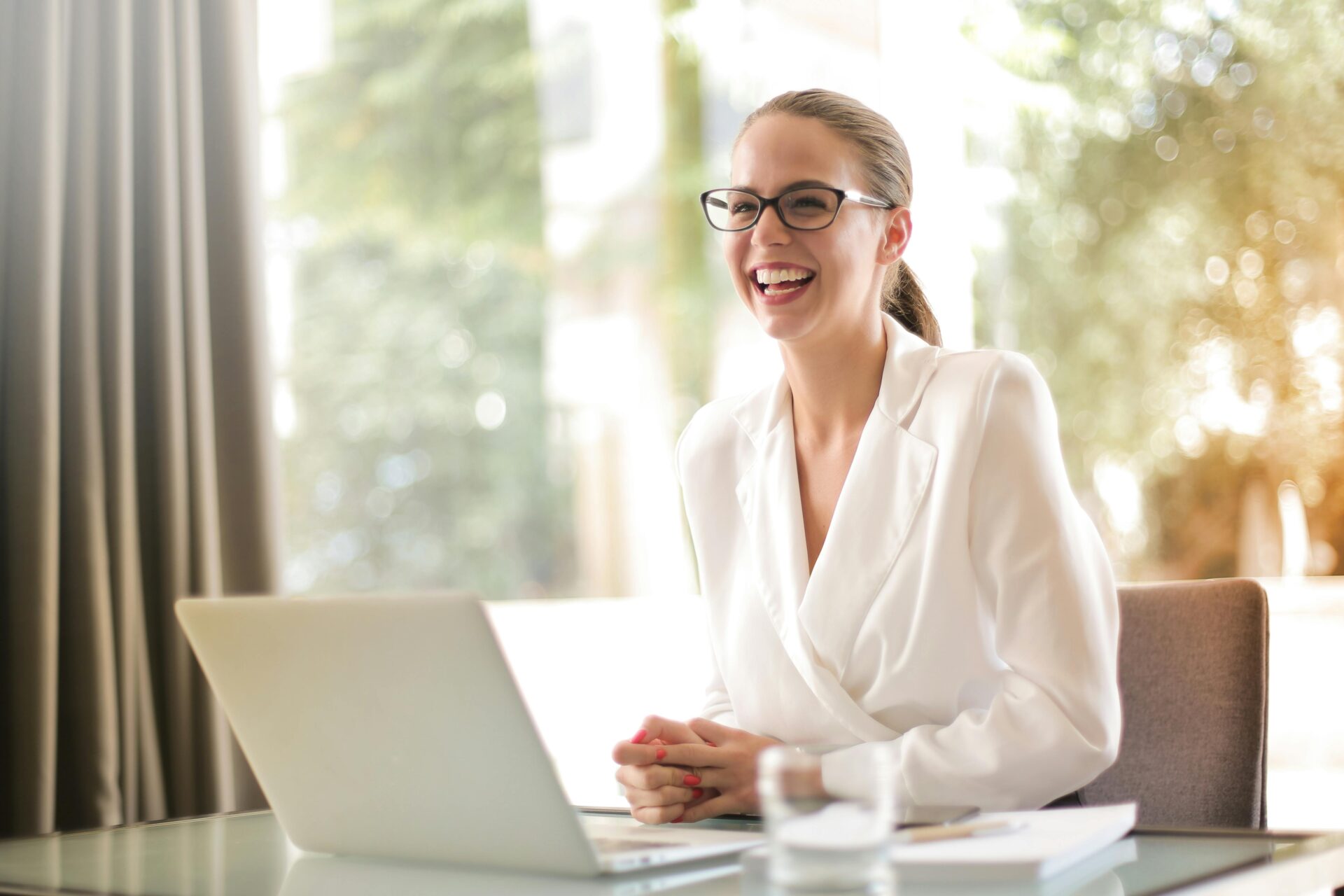 Smiling businesswoman in white blazer working on a laptop in a bright office, representing the concept of what are long-tail keywords in digital marketing.